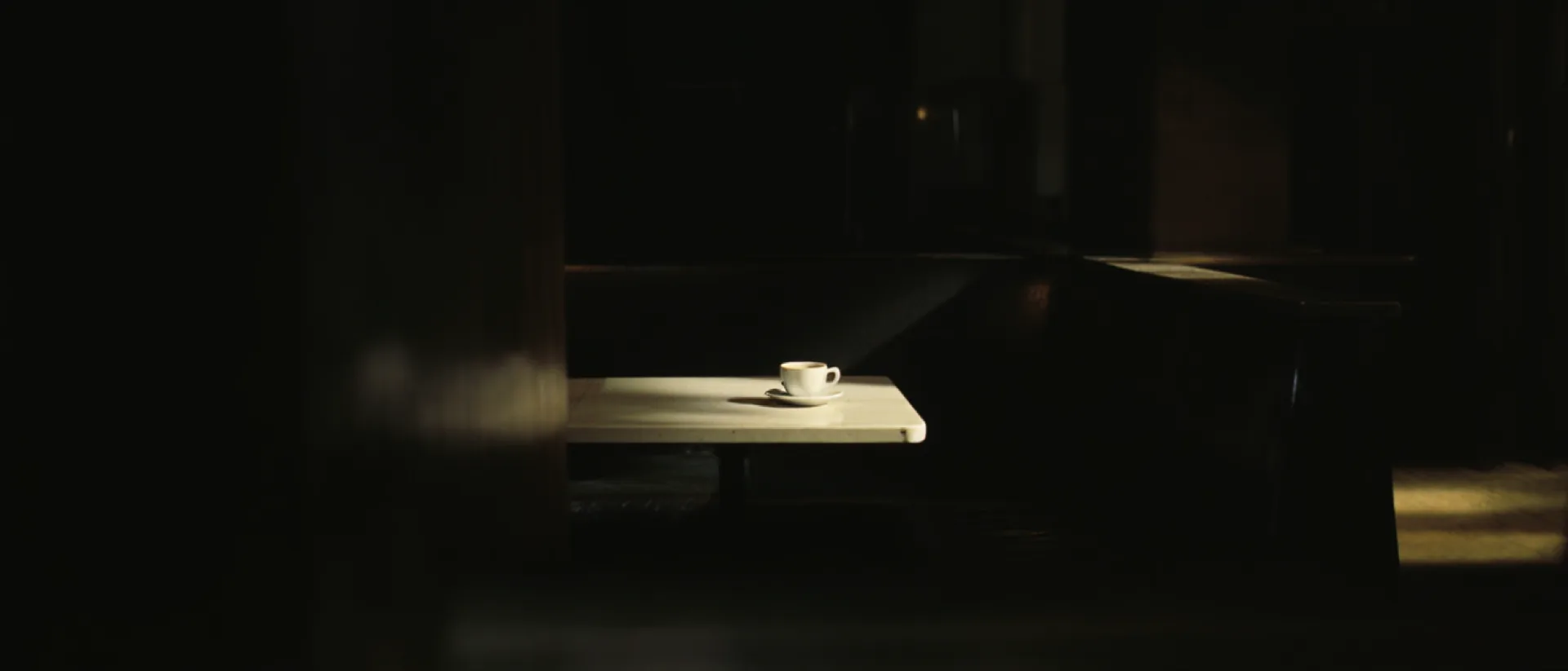A single pale porcelain cup standing in a pool of warm light on a dark walnut bar